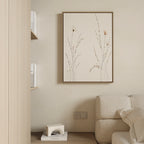 A minimalist living room corner features a beige sofa, the “Prairie Whispers Canvas Art - Vertical” with delicate grasses on the wall, shelves of white books, and a small geometric decor piece for an atmosphere of natural tranquility.