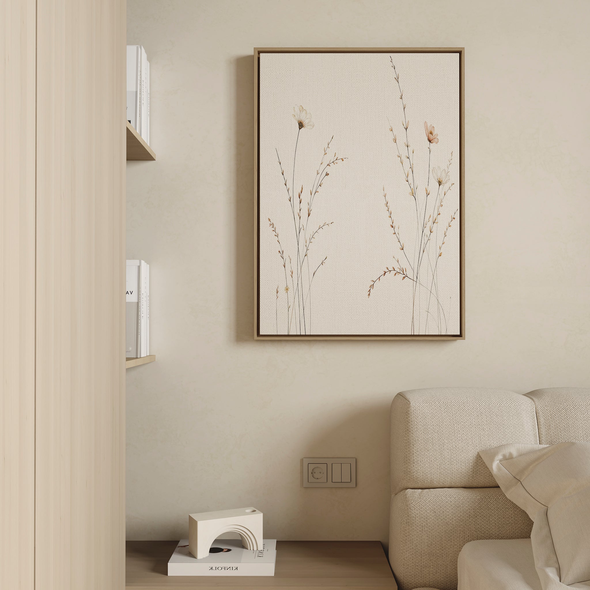A minimalist living room corner features a beige sofa, the “Prairie Whispers Canvas Art - Vertical” with delicate grasses on the wall, shelves of white books, and a small geometric decor piece for an atmosphere of natural tranquility.