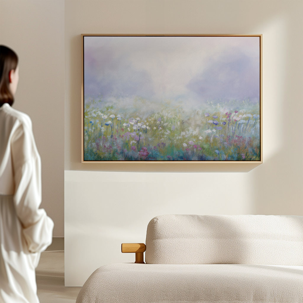 A person in a white robe admires the Purple Haze Canvas Art - Horizontal, which features a misty wildflower meadow scene, mounted on the wall of a softly lit, minimalist living room with a light-colored sofa—ideal decor.