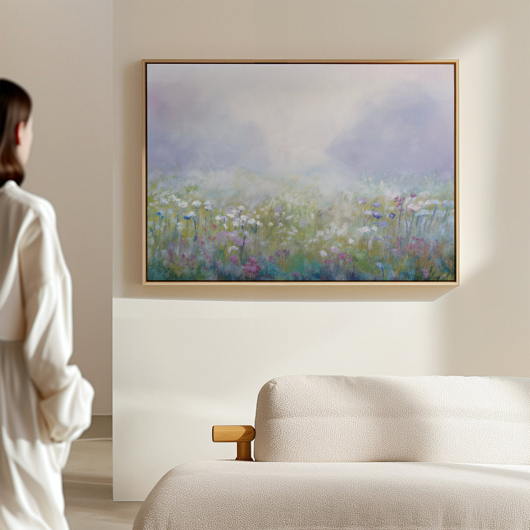 A person in a white robe admires the Purple Haze Canvas Art - Horizontal, which features a misty wildflower meadow scene, mounted on the wall of a softly lit, minimalist living room with a light-colored sofa—ideal decor.