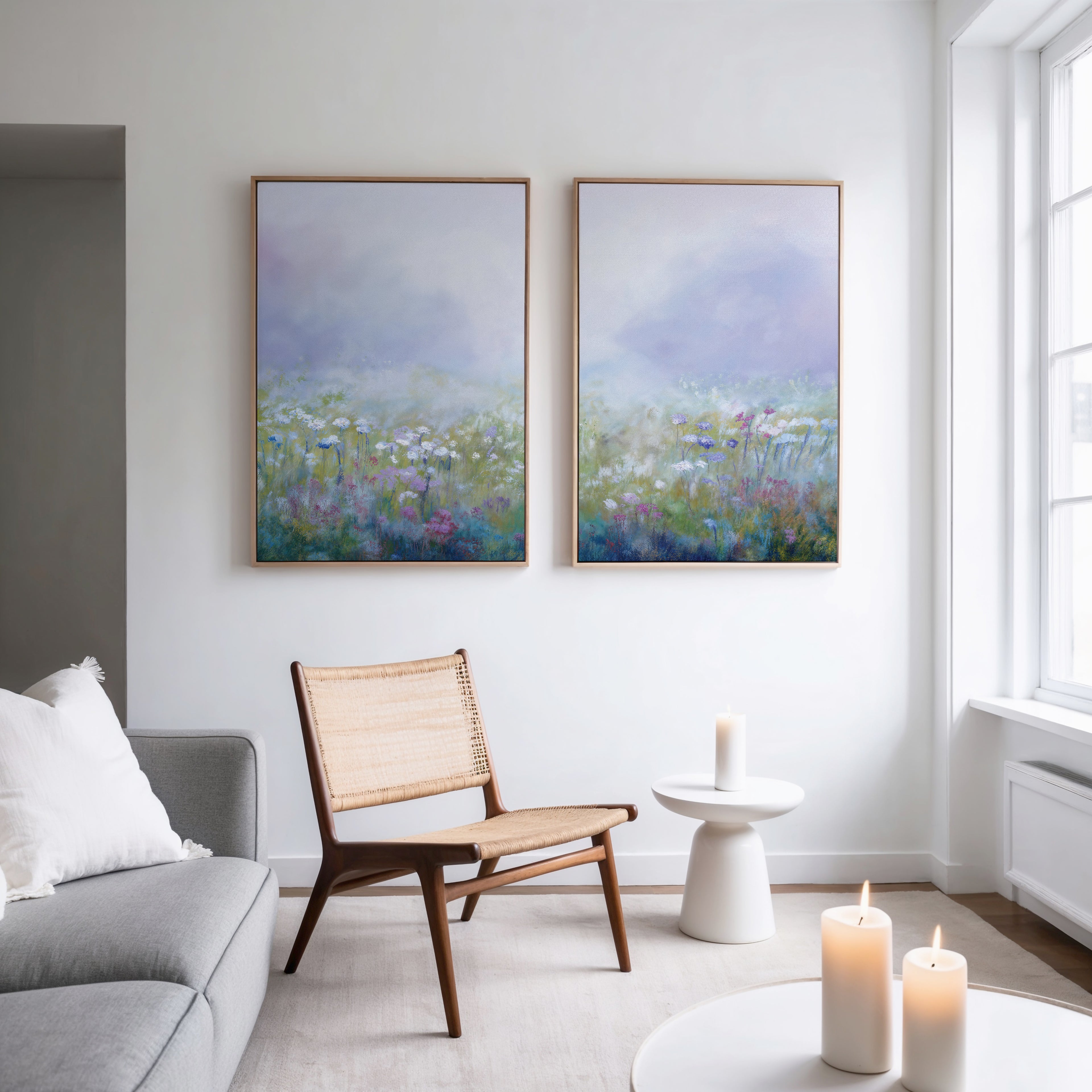 A modern, minimal living room has a gray sofa, woven chair, small white table, three lit candles, and the "Purple Haze - Set of 2" framed canvas prints on a white wall near a large window.