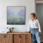 A woman in a white blouse and jeans leans on a wooden cabinet, admiring the Purple Haze Canvas Art - Square above her. A potted plant and book rest on the cabinet, making for inviting living room decor.