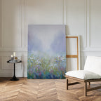 The Purple Haze Canvas Art - Vertical, featuring a misty wildflower field, leans against a white paneled wall in a bright room. A blank frame, cushioned chair, and candlelit table complete this cozy living space on chevron wood floors.