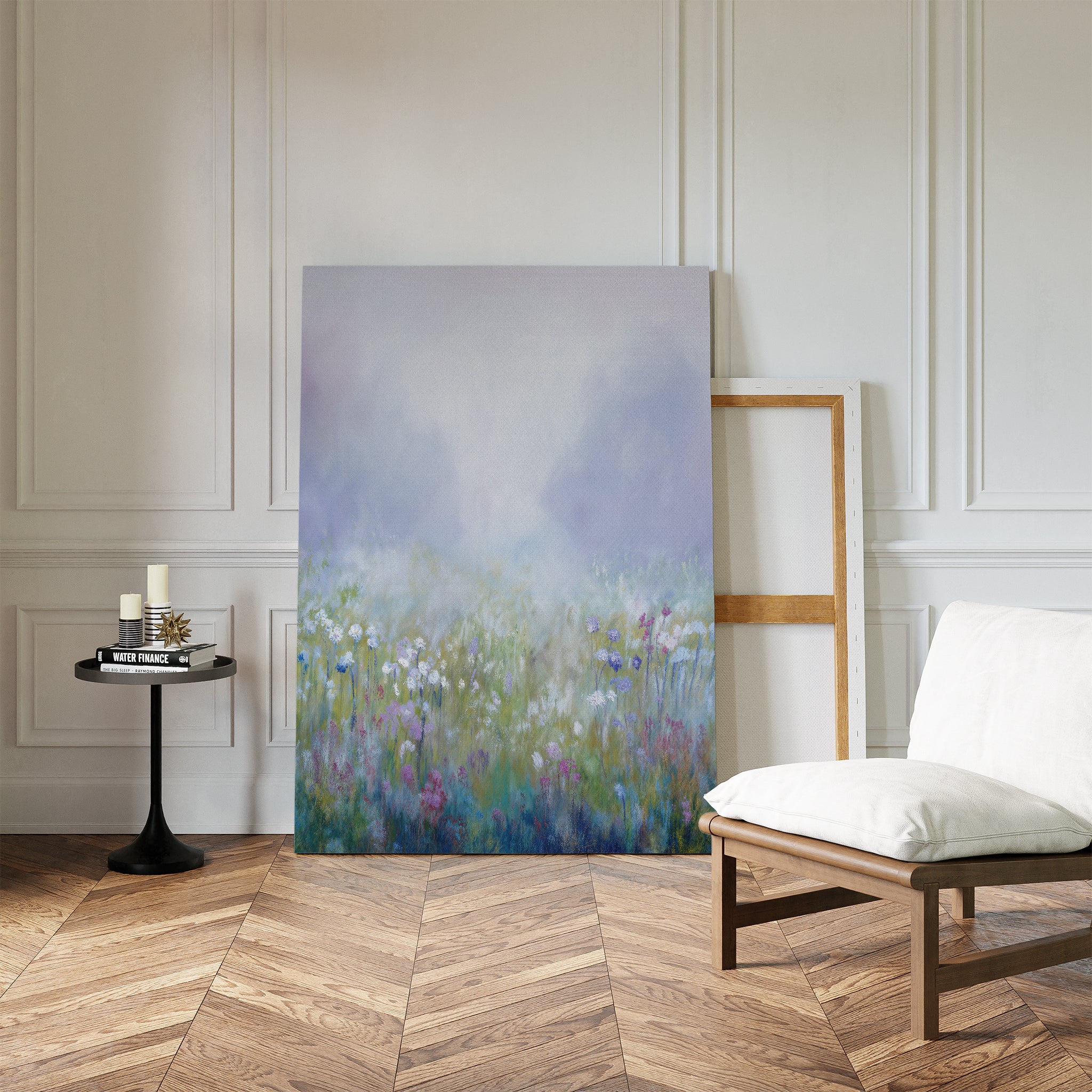 The Purple Haze Canvas Art - Vertical, featuring a misty wildflower field, leans against a white paneled wall in a bright room. A blank frame, cushioned chair, and candlelit table complete this cozy living space on chevron wood floors.