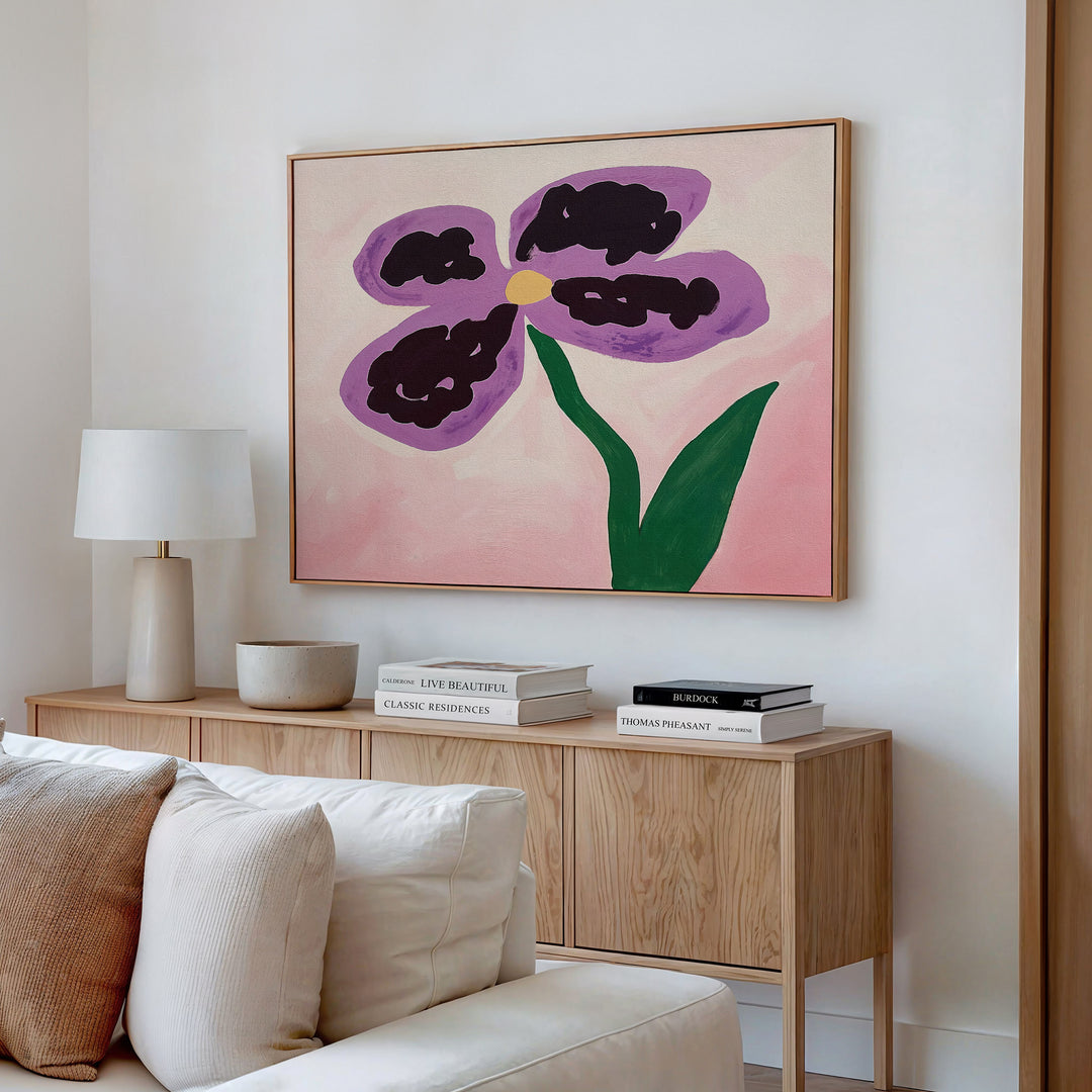 A modern living room with a light wood console, stacked books, a white lamp, a bowl, and the Purple Pansy Canvas Art - Horizontal featuring a vibrant purple flower with green stem on pink background hanging above.
