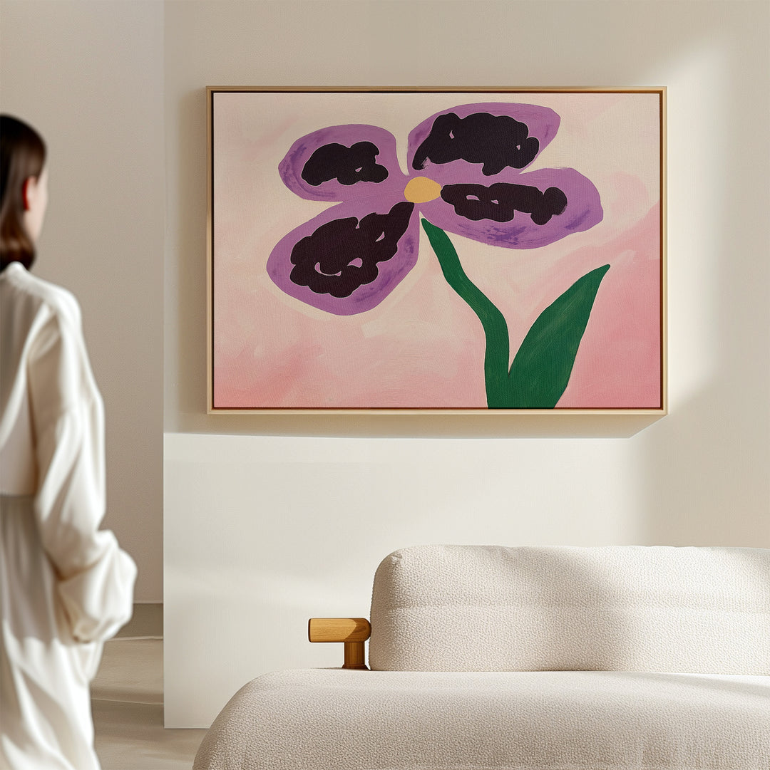 In a softly lit room, a person admires the Purple Pansy Canvas Art - Horizontal, featuring a stylized purple and black flower with a yellow center and green stem on pink, hanging above a beige sofa.