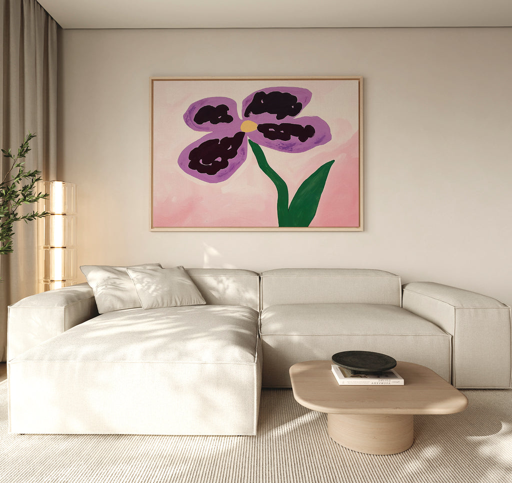 A modern living room with a light beige sectional, light wood coffee table, and the Purple Pansy Canvas Art - Horizontal featuring a purple flower on a pink background hanging above the sofa.