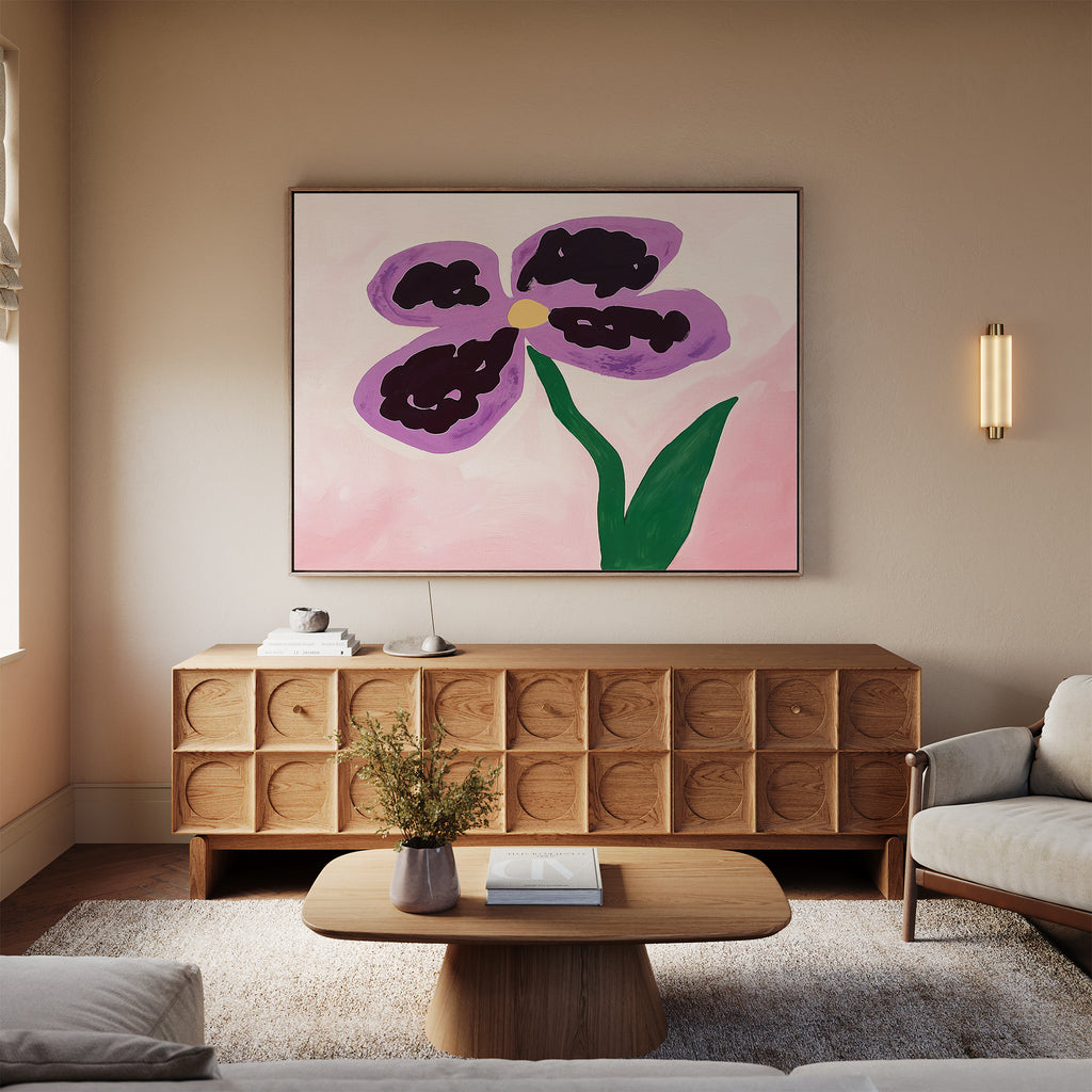 A modern living room with a light sofa, wooden coffee table, and sideboard features the Purple Pansy Canvas Art - Horizontal above the sideboard—a vivid botanical print of a purple-petaled flower with a green stem.