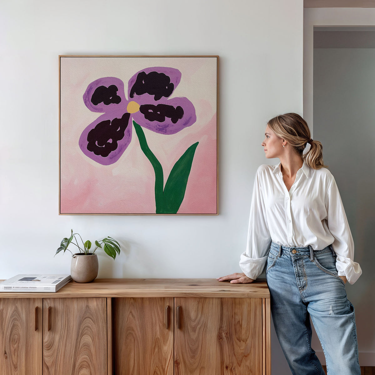 A woman in a white blouse and jeans leans against a wooden cabinet, admiring the Purple Pansy Canvas Art - Square on a white wall. A small potted plant and books complete the inviting, Nordic-inspired look.