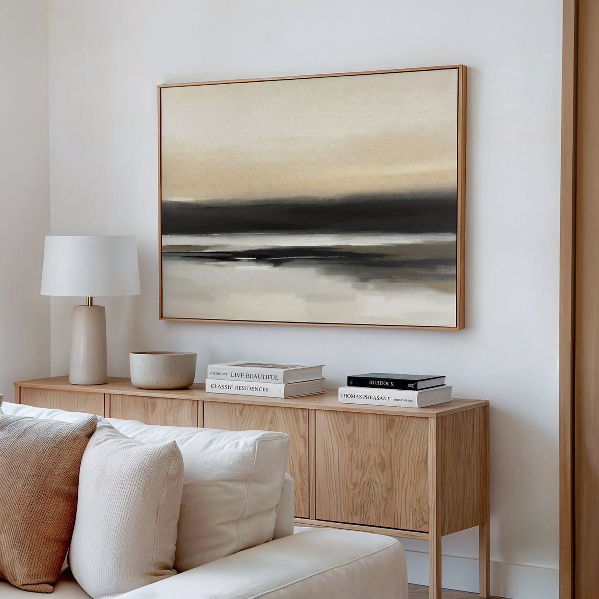 A light, modern living room features a wooden sideboard with books, a lamp, and decor bowl. Above hangs the Quiet Shoreline Canvas Art - Horizontal in serene neutral tones. A white sofa sits in this calming space.