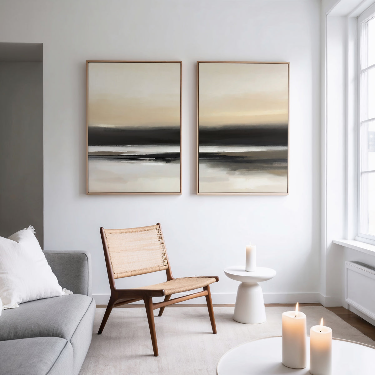 A modern, minimal living room features a gray sofa, woven chair, small round white table with three lit candles, and the Quiet Shoreline - Set Of 2 wall art pieces on a white wall by a large window.