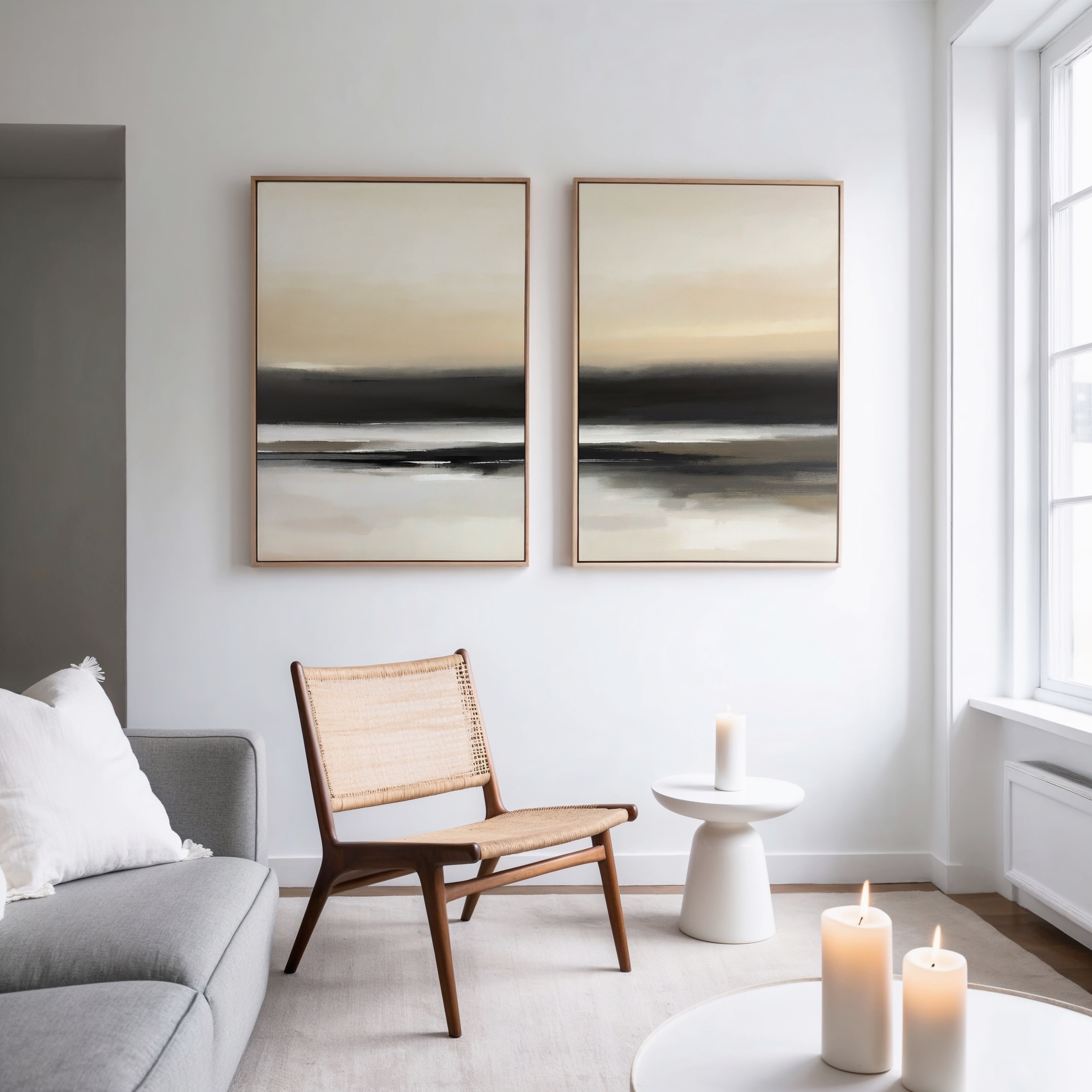 A modern, minimal living room features a gray sofa, woven chair, small round white table with three lit candles, and the Quiet Shoreline - Set Of 2 wall art pieces on a white wall by a large window.