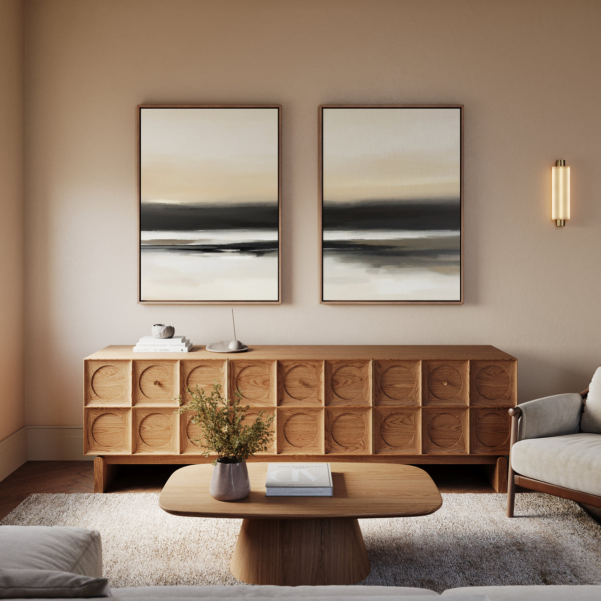 A modern living room features a wooden sideboard, the "Quiet Shoreline - Set Of 2" minimalist abstract landscape wall art, a round coffee table with books and a vase with greenery, a light gray armchair, and soft neutral walls.