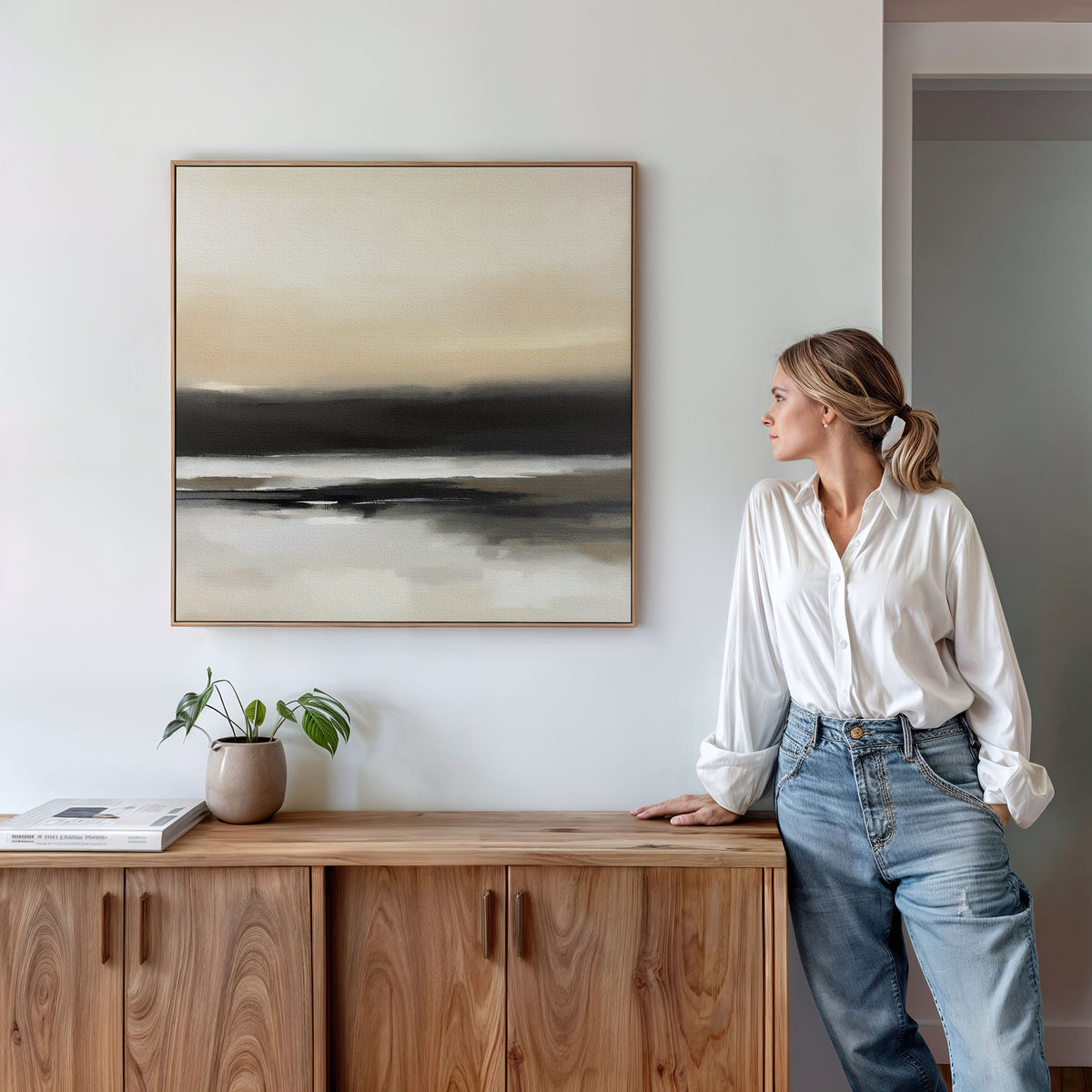 A woman in a white blouse and jeans leans on a wooden cabinet, admiring the Quiet Shoreline Canvas Art - Square with neutral tones above her. Minimalist decor includes a small potted plant and a book neatly placed on the cabinet.