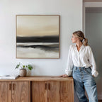 A woman in a white blouse and jeans leans on a wooden cabinet, admiring the Quiet Shoreline Canvas Art - Square with neutral tones above her. Minimalist decor includes a small potted plant and a book neatly placed on the cabinet.