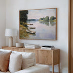 A cozy living room corner features a wooden sideboard, books, a lamp, and the serene River Tranquil Canvas Art - Horizontal depicting boats on a riverbank surrounded by trees hanging above.