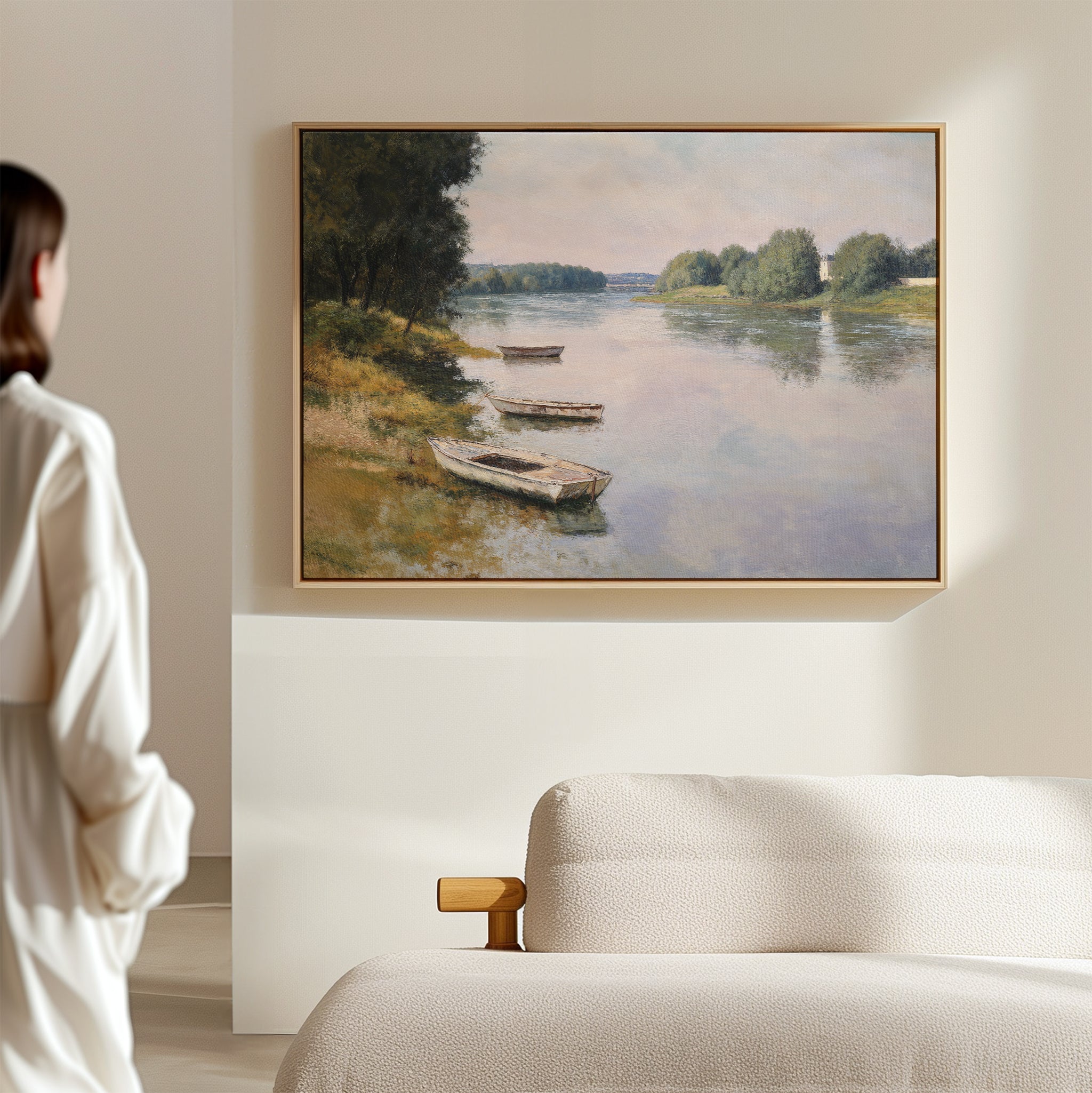 A person admires the River Tranquil Canvas Art - Horizontal, depicting boats on a calm river with green trees, mounted above a modern upholstered sofa in a softly lit room.