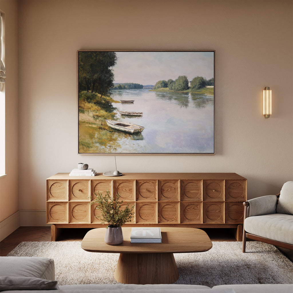 A serene living room features a light wooden sideboard with the River Tranquil Canvas Art - Horizontal above it, a coffee table holding a vase and book, a beige sofa, soft rug, and warm lighting from a wall sconce.