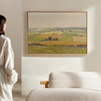 A person in white stands by a beige sofa, admiring the Rolling Fields Canvas Art - Horizontal—a framed landscape of green and yellow fields—displayed on a light-colored wall.