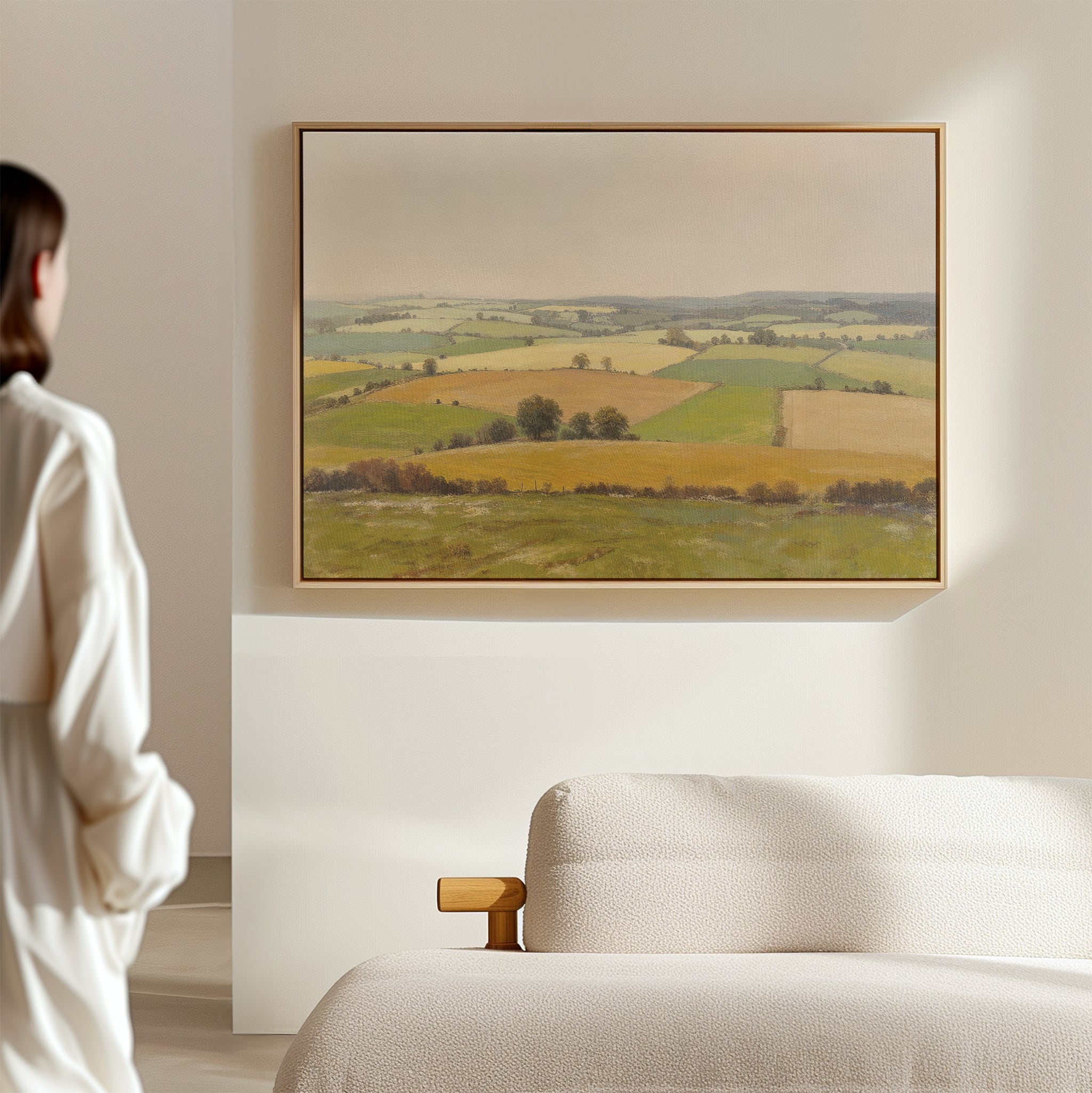 A person in white stands by a beige sofa, admiring the Rolling Fields Canvas Art - Horizontal—a framed landscape of green and yellow fields—displayed on a light-colored wall.