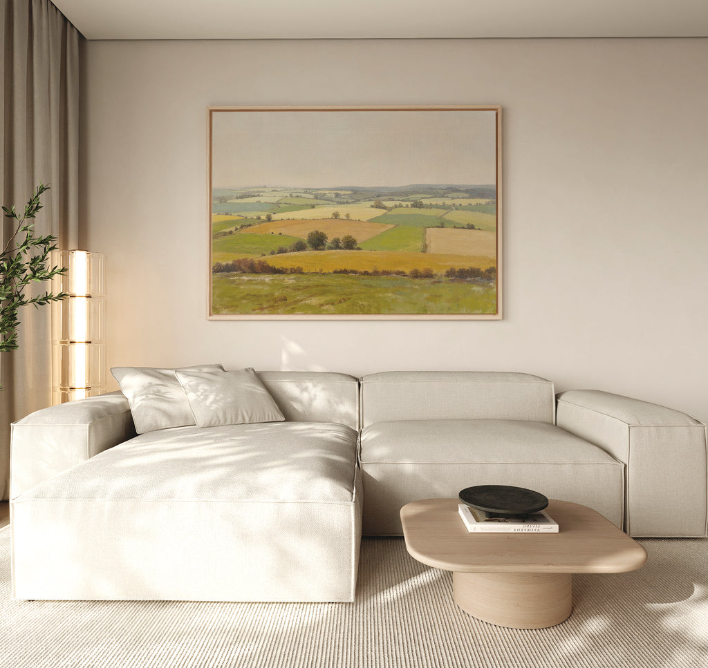 A modern living room with a light beige sectional sofa, low wood coffee table with a black bowl, tall plant, and serene wall art—a large Rolling Fields Canvas Art - Horizontal—displayed above the sofa.