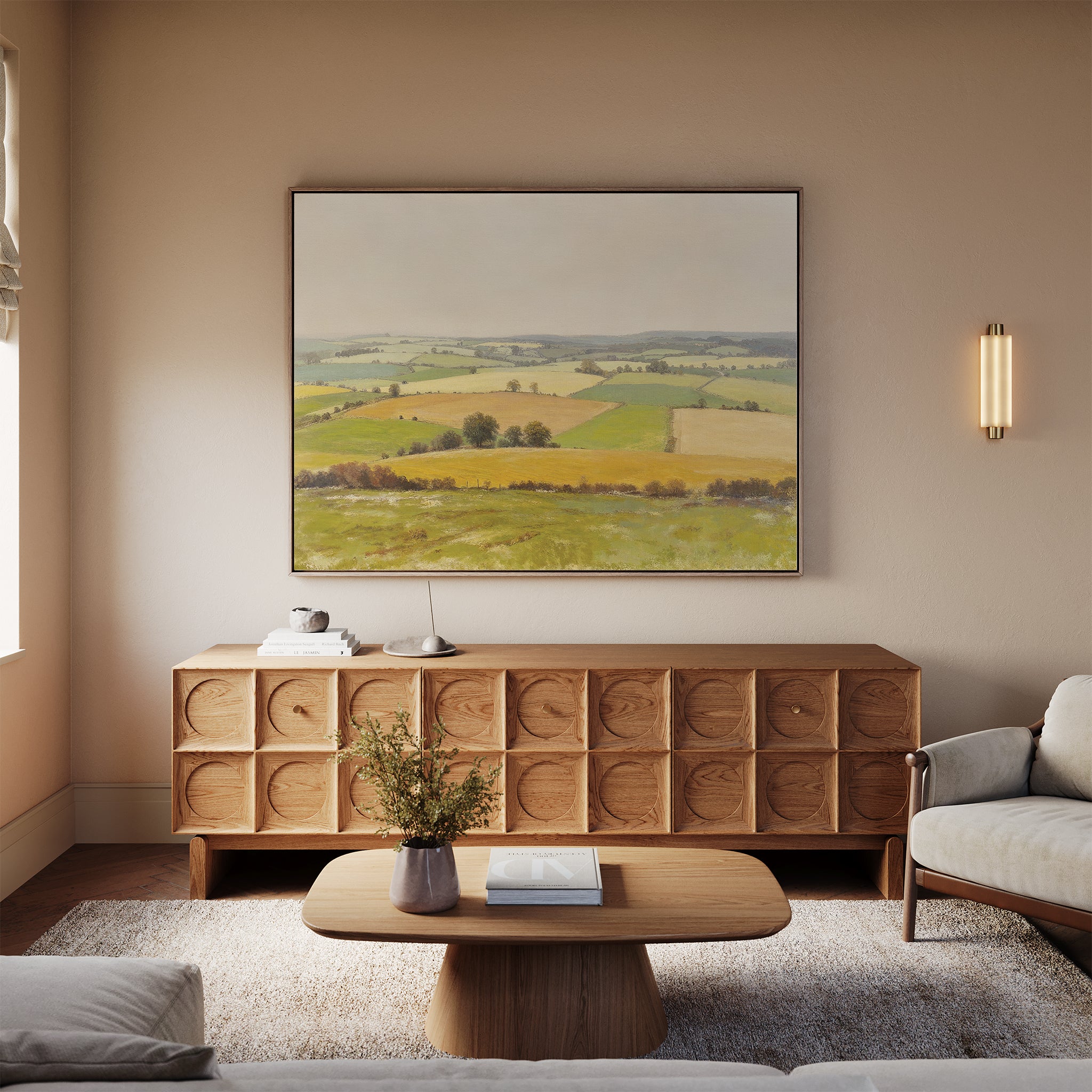 A cozy living room features a beige sofa, wooden coffee table, and sideboard. Above the sideboard hangs the Rolling Fields Canvas Art - Horizontal, depicting green and yellow fields. Soft lighting adds a warm, inviting glow.