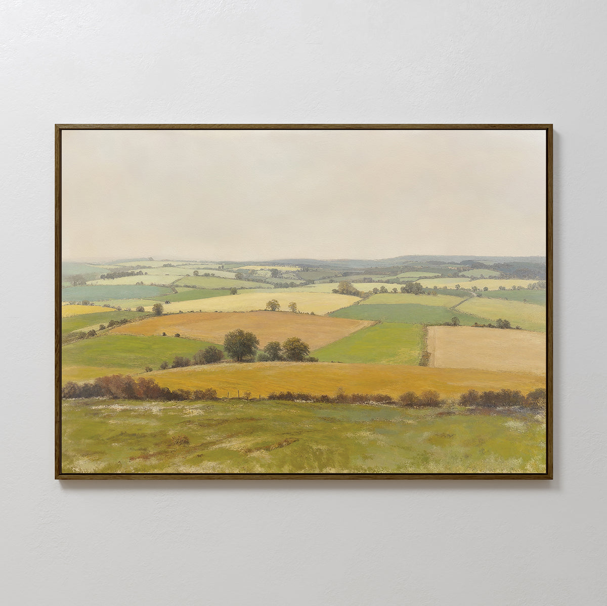 The Rolling Fields Canvas Art - Horizontal displays a pastoral countryside with green and yellow fields, scattered trees, and a hazy sky, elegantly framed and hung on a plain light-colored wall.