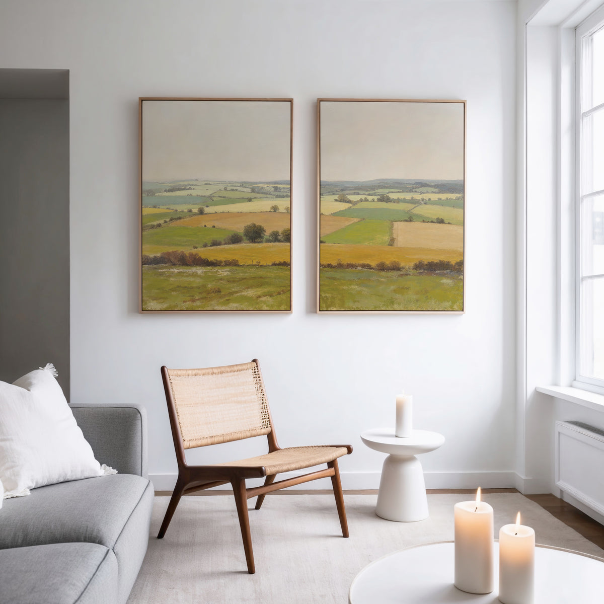 A modern living room with a gray sofa, wooden chair, and small round white table with a lit candle. Above, the "Rolling Fields - Set of 2" canvas prints bring tranquil countryside charm as sunlight streams in through large windows.
