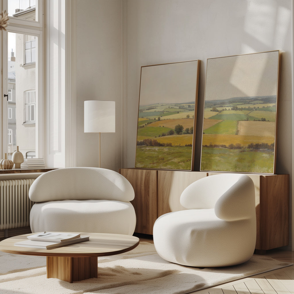 A bright, modern living room with two rounded white chairs, a low wooden table, and Rolling Fields - Set of 2 countryside canvas prints on a wooden sideboard against the wall near tall windows.