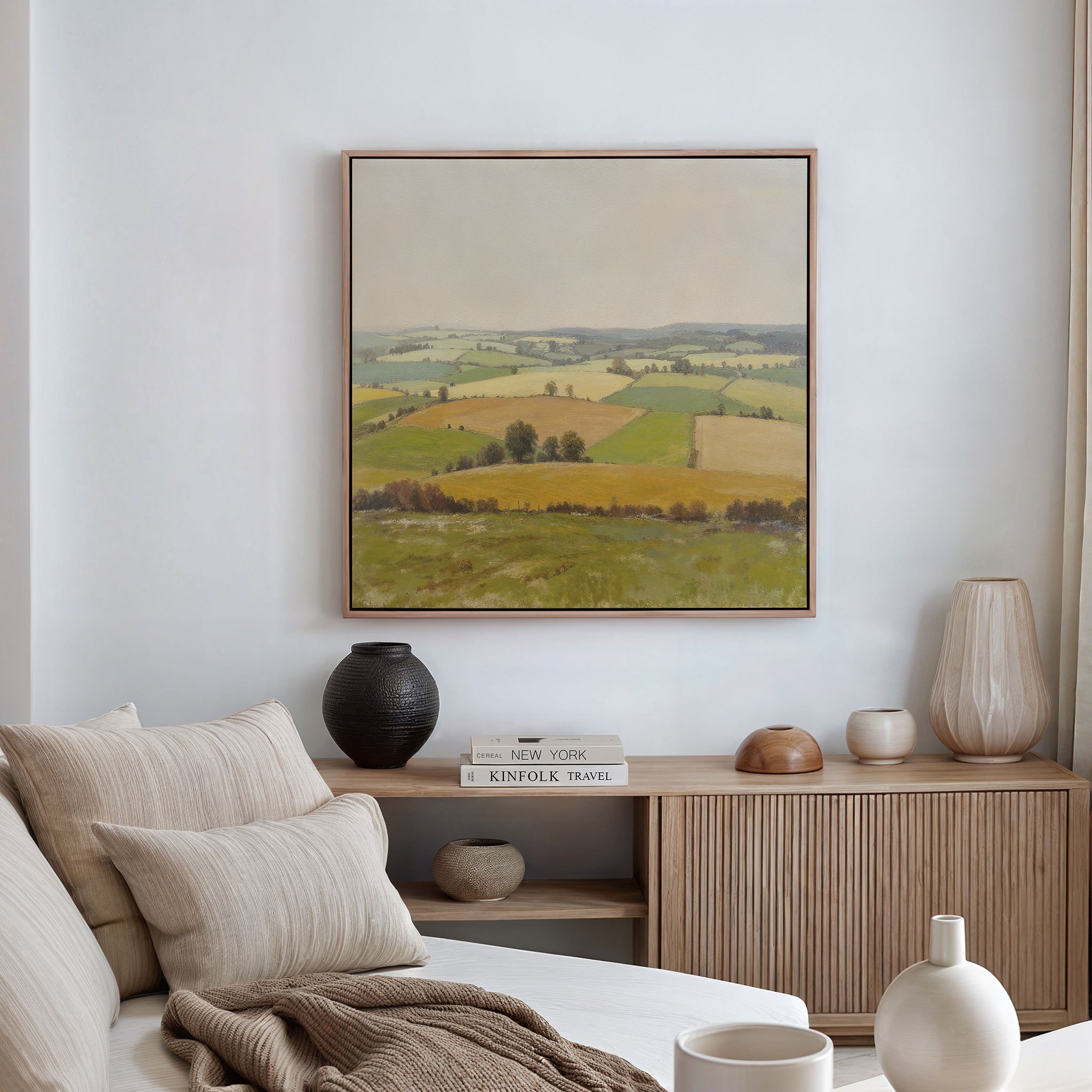 A cozy living room features a beige sofa, textured pillows, wooden sideboard, decorative vases, and the Rolling Fields Canvas Art - Square—a landscape painting of green and yellow fields—bringing countryside charm to a white wall.