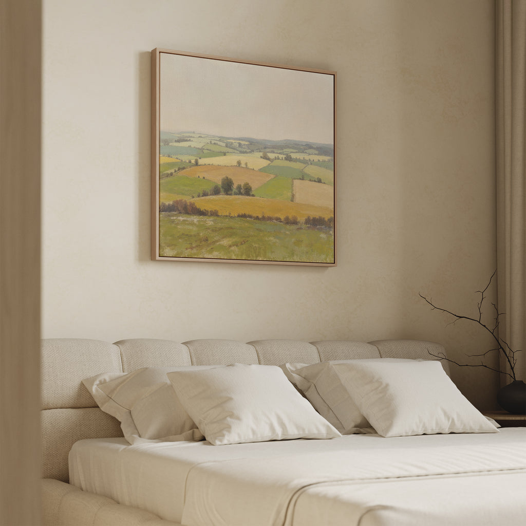 A cozy bedroom features a beige bed with white pillows and the Rolling Fields Canvas Art - Square, displaying soft green and yellow field scenes above the headboard. A simple vase with bare branches rests on the side table.