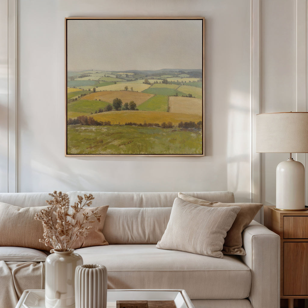 A bright, modern living room features a beige sofa, neutral pillows, and a side table with a cream lamp. Above vases of dried flowers hangs the Rolling Fields Canvas Art - Square, bringing tranquility with its pastoral countryside scene.