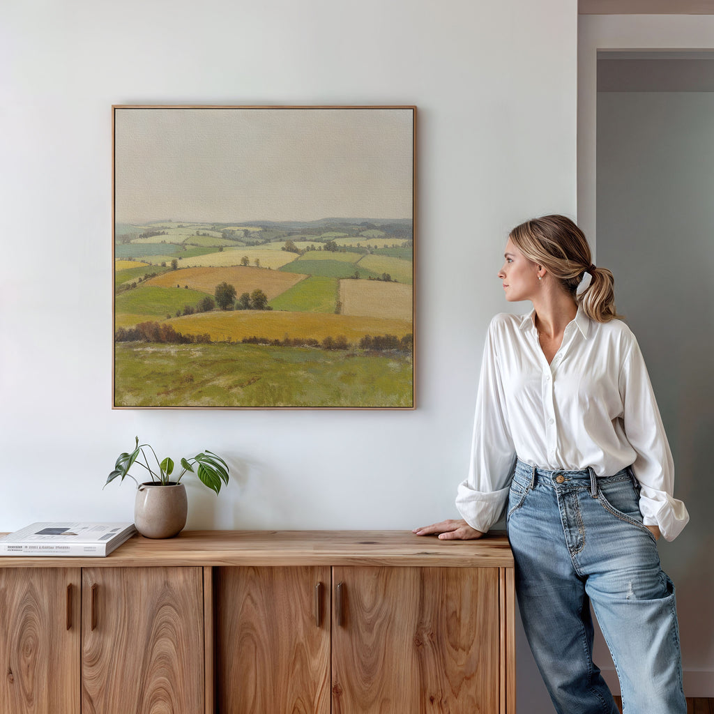 A woman in a white blouse and jeans stands by a wooden cabinet, gazing at serene wall art—a framed Rolling Fields Canvas Art - Square depicting pastoral countryside. A potted plant and books sit atop the cabinet.