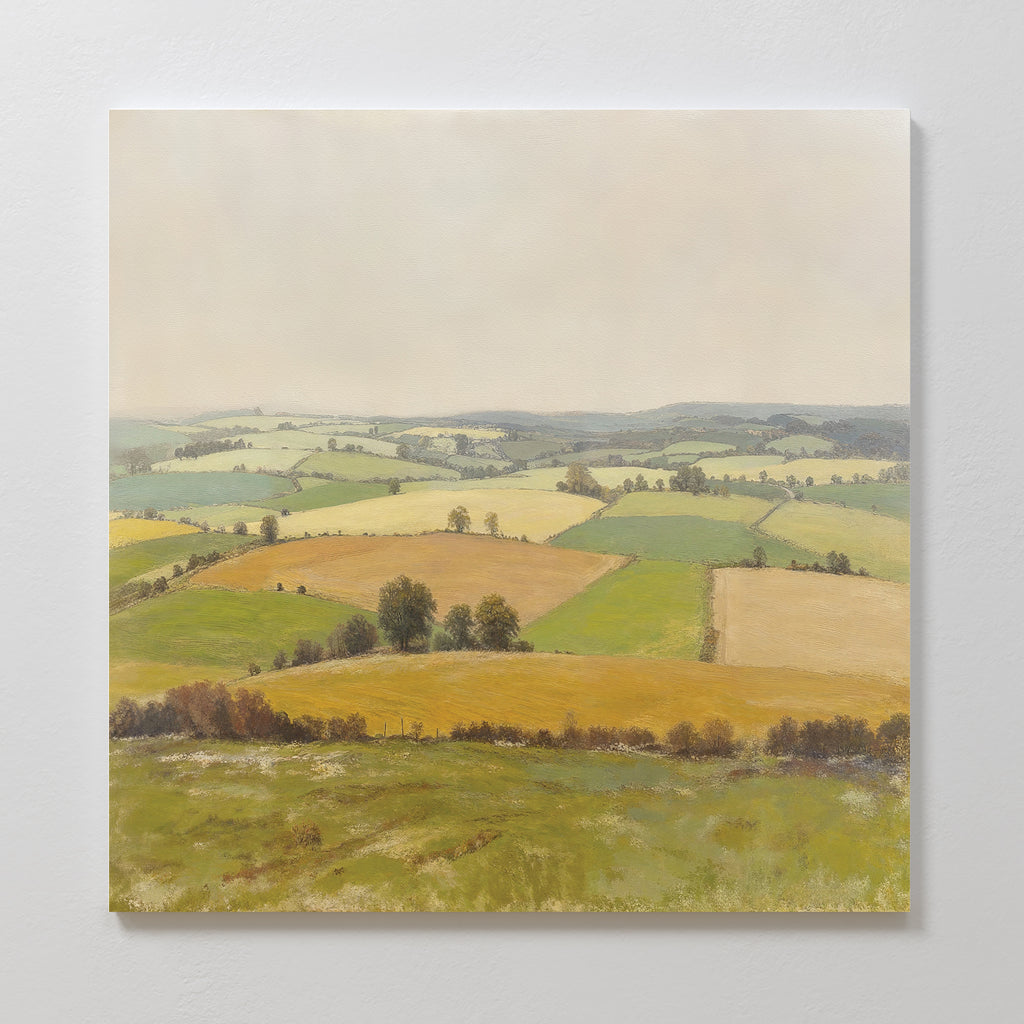 The Rolling Fields Canvas Art - Square showcases a rural landscape with green, yellow, and brown patchwork fields, rolling hills, scattered trees, and a pale sky. This serene wall art is displayed on a white wall.