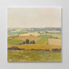 The Rolling Fields Canvas Art - Square showcases a rural landscape with green, yellow, and brown patchwork fields, rolling hills, scattered trees, and a pale sky. This serene wall art is displayed on a white wall.