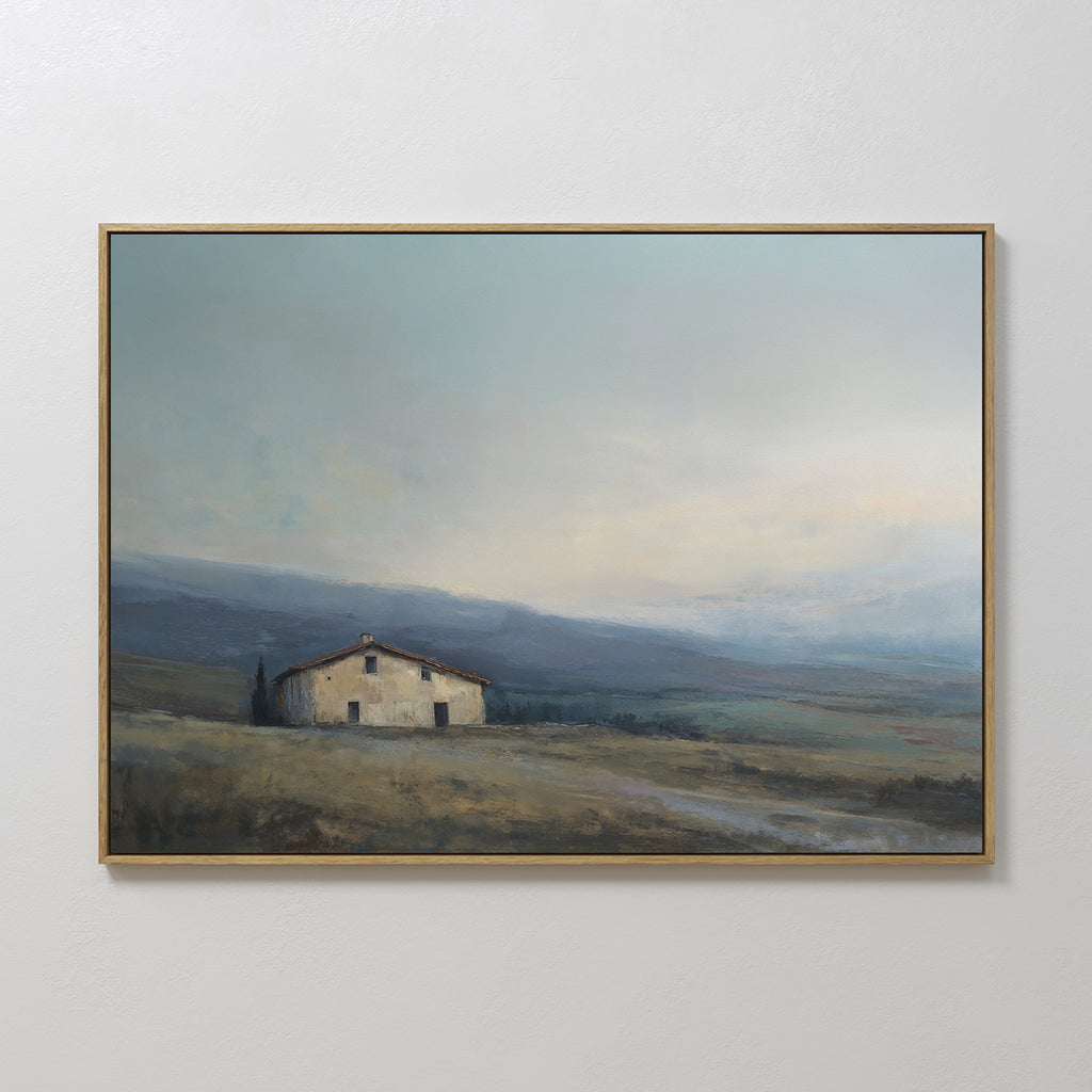 The Rustic Solitude Canvas Art - Horizontal showcases a small, rustic house in a grassy field with rolling hills and a cloudy sky, framed in thin gold and displayed on a light-colored wall for charming farmhouse appeal.