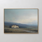 The Rustic Solitude Canvas Art - Horizontal showcases a small, rustic house in a grassy field with rolling hills and a cloudy sky, framed in thin gold and displayed on a light-colored wall for charming farmhouse appeal.