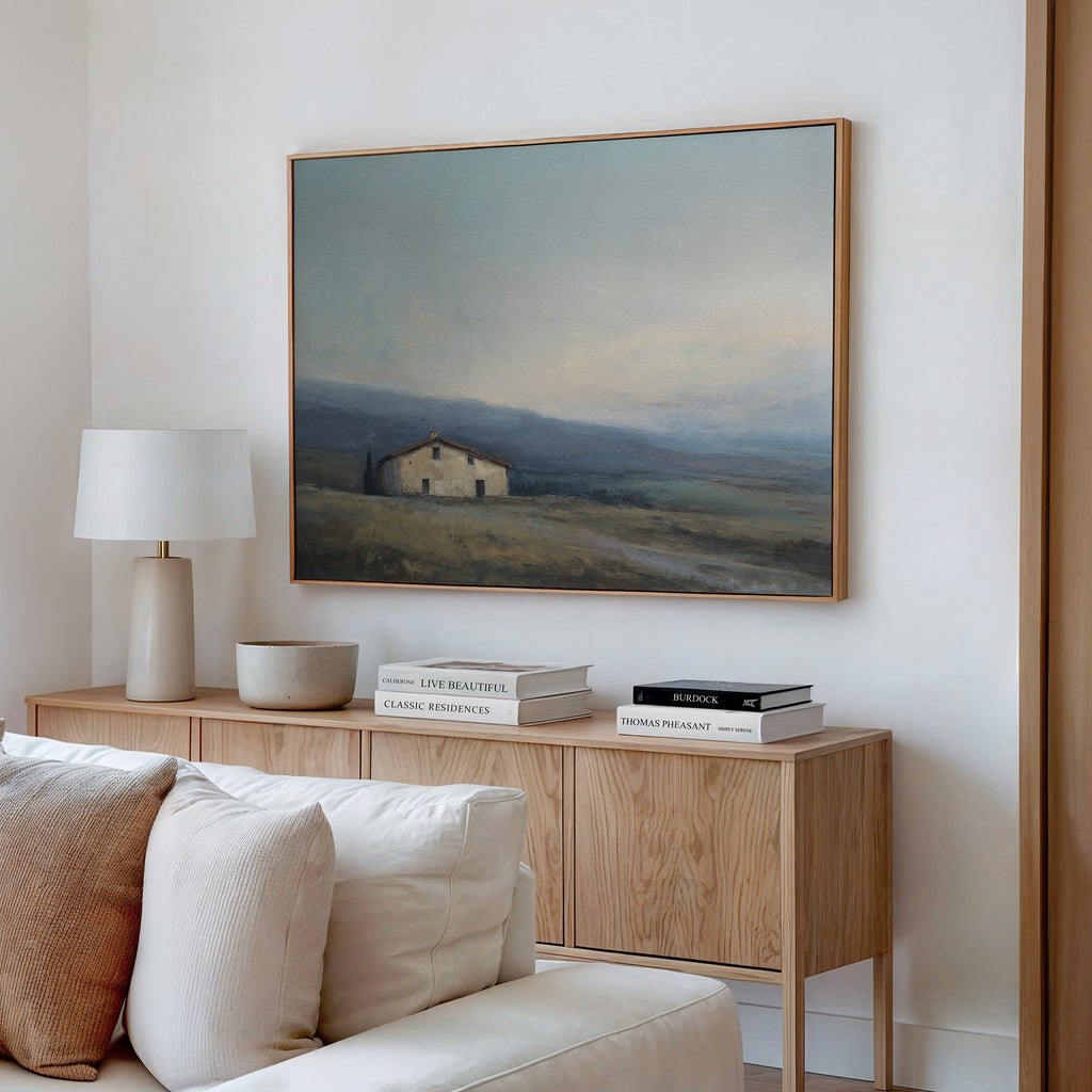 A cozy living room features a wooden sideboard with books and decor, a white lamp, and the Rustic Solitude Canvas Art - Horizontal—a landscape painting of a house in a misty field—hanging above a beige couch.