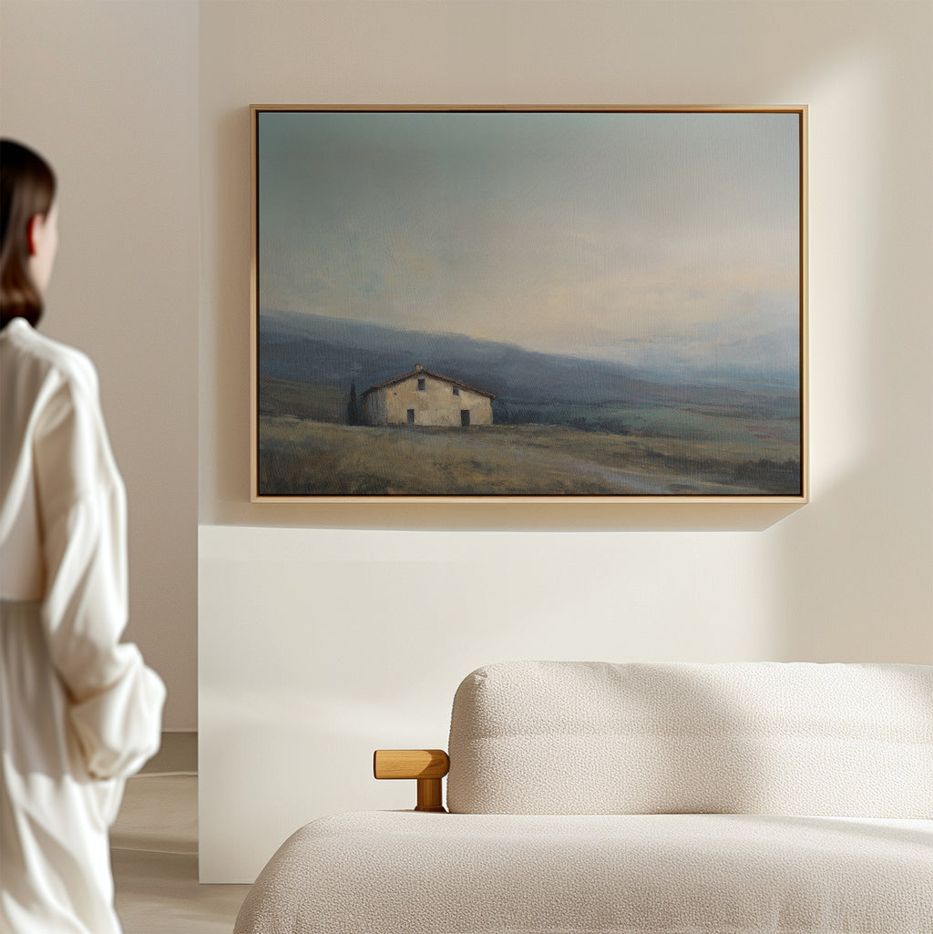 A woman in white stands in a minimal, sunlit room, admiring the Rustic Solitude Canvas Art - Horizontal, an elegant landscape of a countryside house in a field, displayed above the cream-colored sofa.