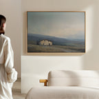 A woman in white stands in a minimal, sunlit room, admiring the Rustic Solitude Canvas Art - Horizontal, an elegant landscape of a countryside house in a field, displayed above the cream-colored sofa.