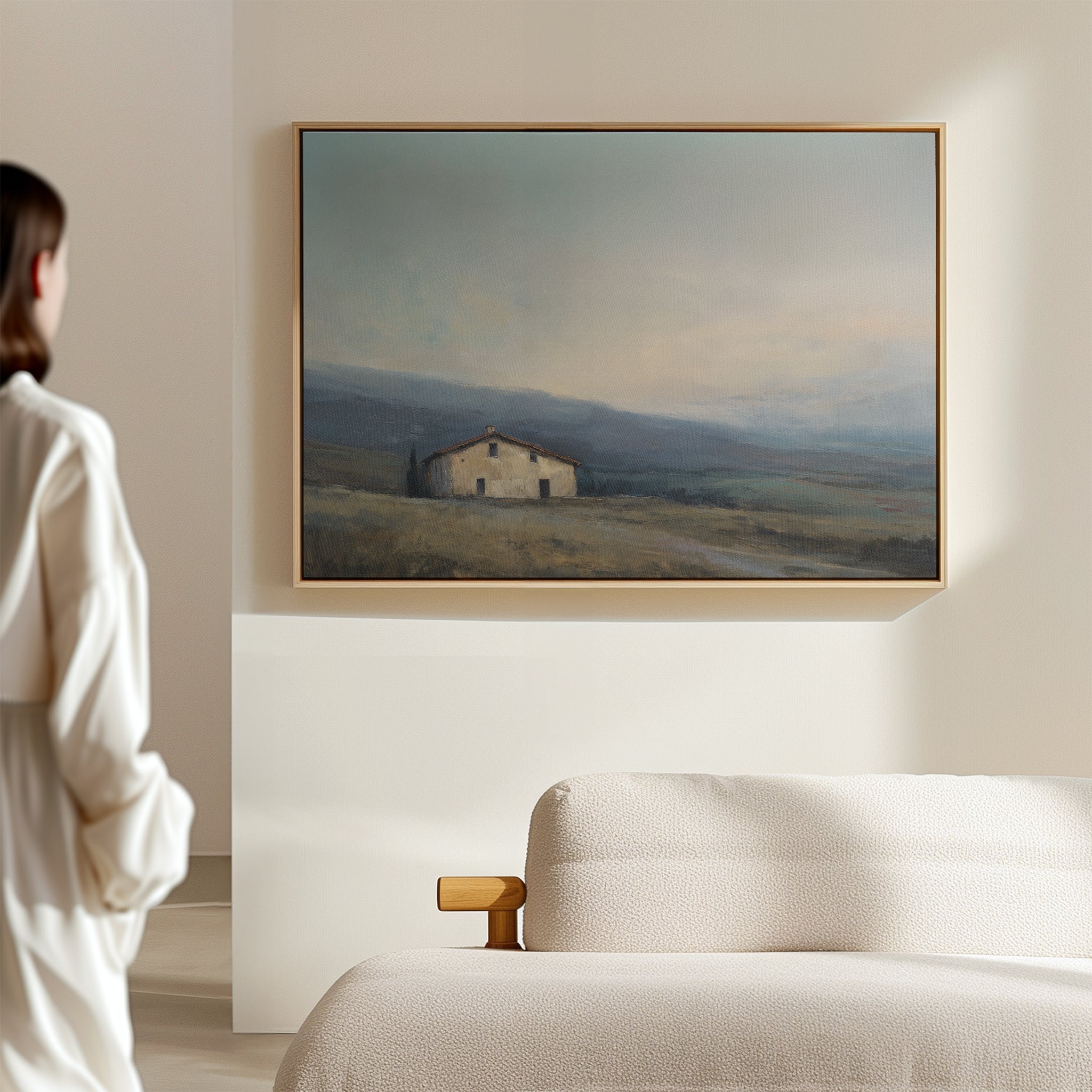 A woman in white stands in a minimal, sunlit room, admiring the Rustic Solitude Canvas Art - Horizontal, an elegant landscape of a countryside house in a field, displayed above the cream-colored sofa.