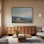 A cozy living room features a light wood sideboard with the Rustic Solitude Canvas Art - Horizontal above, a light sofa, neutral walls softly lit by a wall sconce, and a small coffee table with a book and vase.