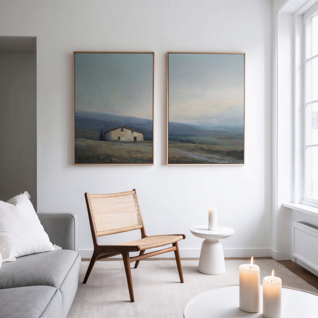 A modern living room with a light gray sofa, wooden chair, and small white table with candles. The space is decorated with "Rustic Solitude - Set of 2" countryside canvas prints hanging above the chair on the white wall.