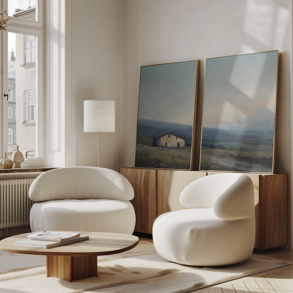 A modern living room features two white rounded armchairs, a low wooden coffee table, a wooden sideboard, and serene landscape decor with the Rustic Solitude - Set of 2 large countryside canvas prints resting against tall windows.