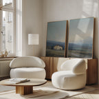 A modern living room features two white rounded armchairs, a low wooden coffee table, a wooden sideboard, and serene landscape decor with the Rustic Solitude - Set of 2 large countryside canvas prints resting against tall windows.