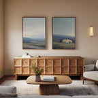 A cozy living room shows a wooden sideboard with geometric patterns, a matching coffee table with decor, light walls, rustic farmhouse wall art, the "Rustic Solitude - Set of 2" landscape paintings, a wall sconce, and a beige armchair with cushions.