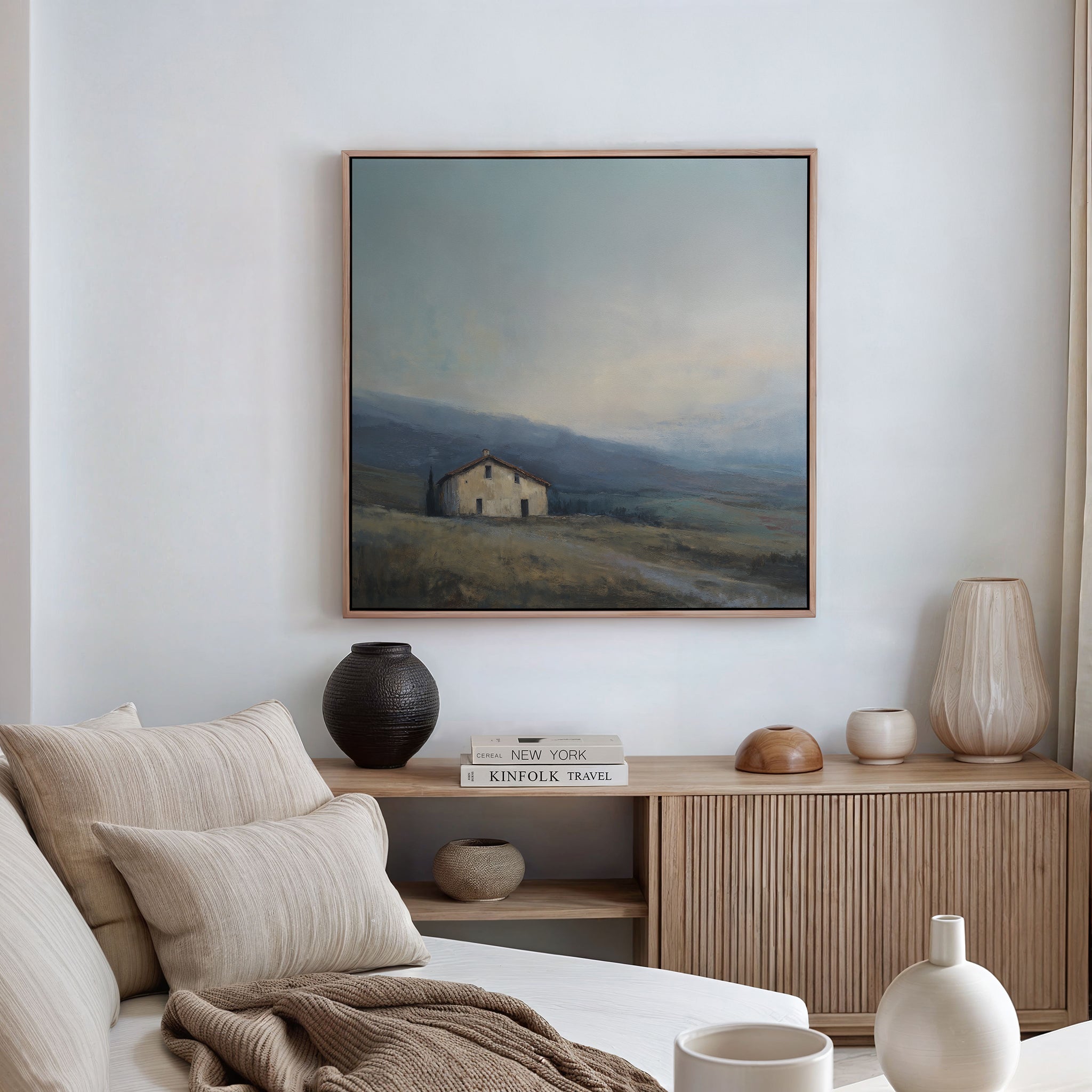 A minimal, neutral-toned living room features a beige sofa with striped pillows, a wooden sideboard with vases and books, and the Rustic Solitude Canvas Art - Square displayed above the mantel.