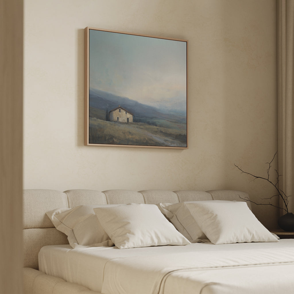 A minimalist bedroom features a beige upholstered bed, four white pillows, and the Rustic Solitude Canvas Art - Square above the headboard. Soft natural light and neutral tones create a calm, serene atmosphere.