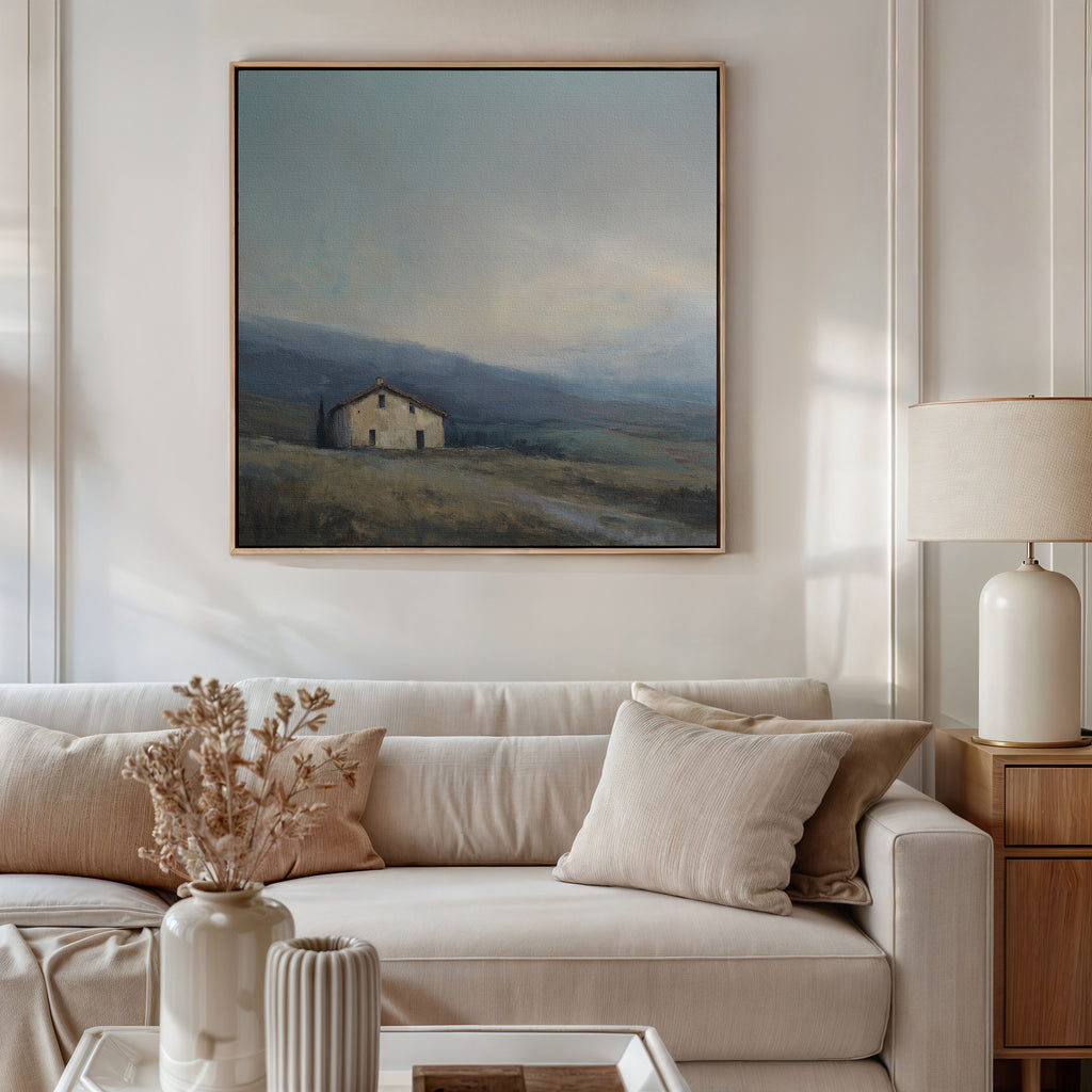 A modern living room featuring a beige sofa, neutral throw pillows, a ceramic vase with dried flowers, and a table lamp. Above the sofa hangs the Rustic Solitude Canvas Art - Square, depicting a serene house in misty countryside.