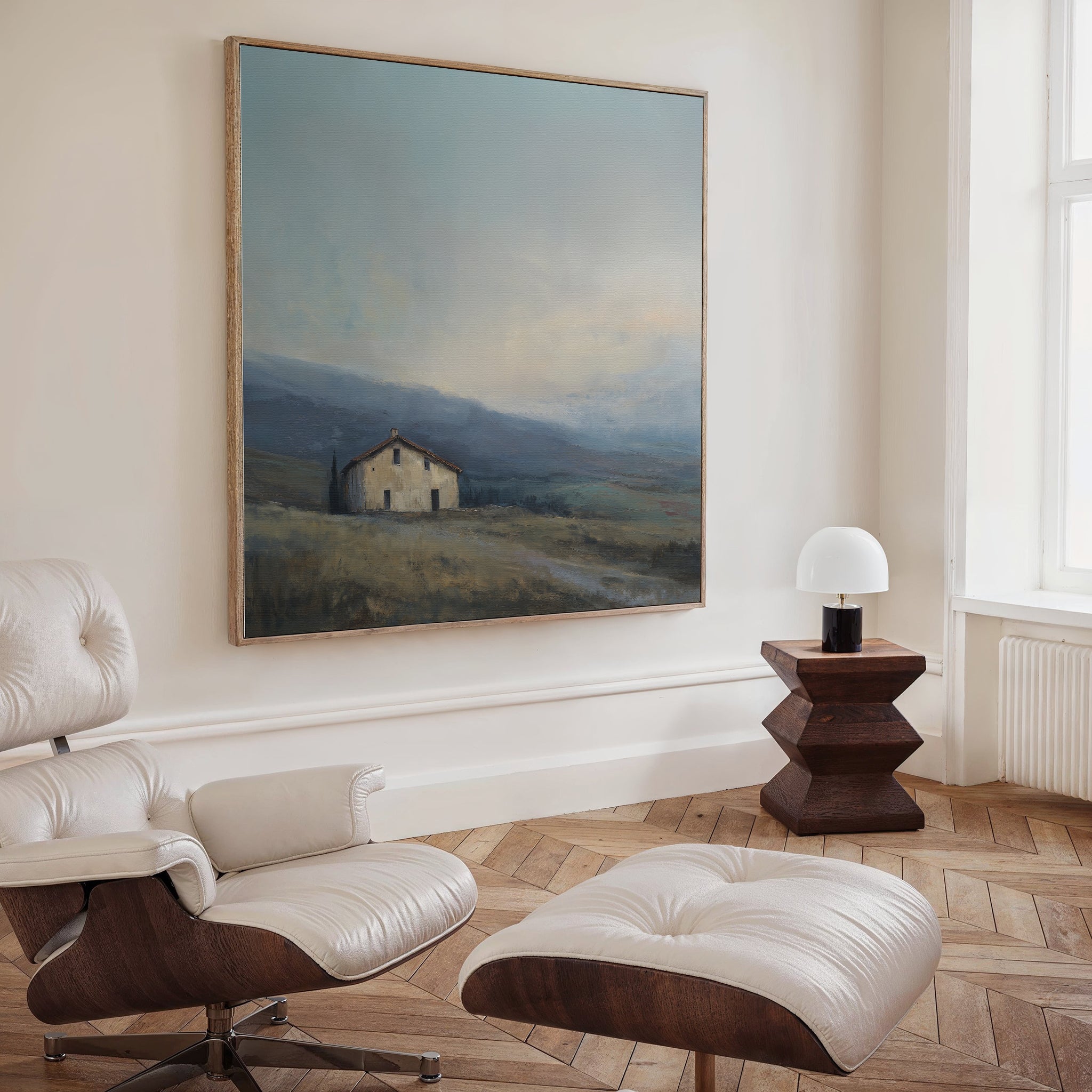 A cozy room with a modern white lounge chair and ottoman, a geometric wooden side table with a white lamp, and the Rustic Solitude Canvas Art - Square displayed above herringbone wood floors.